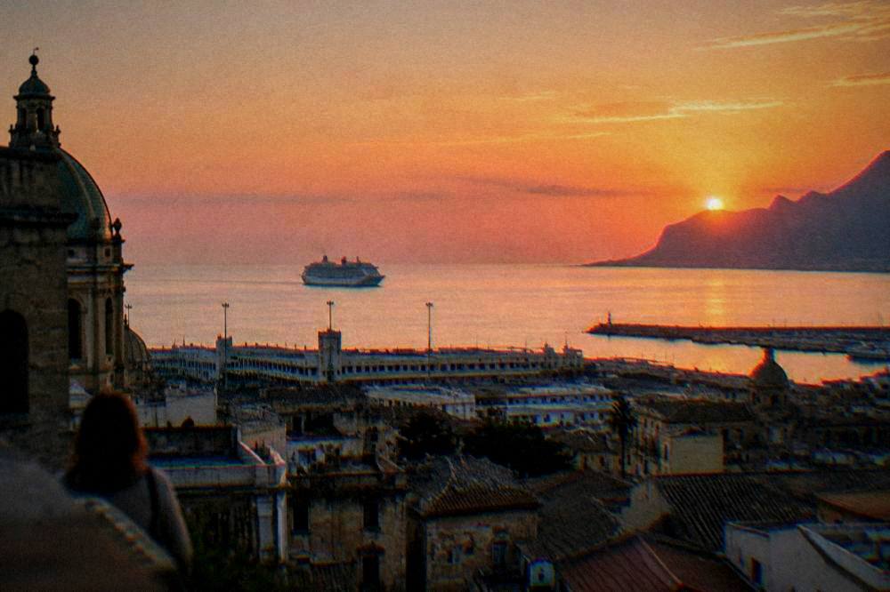 View across Palermo rooftops toward the sea with a ship on the horizon at sunset