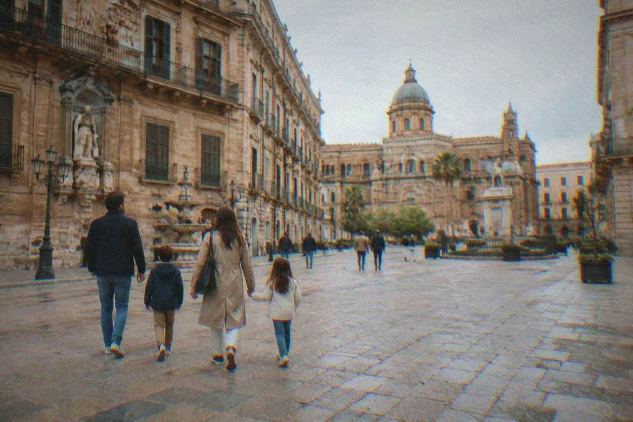 A family with two children walking across a sunlit square lined with old ornate buildings in Palermo