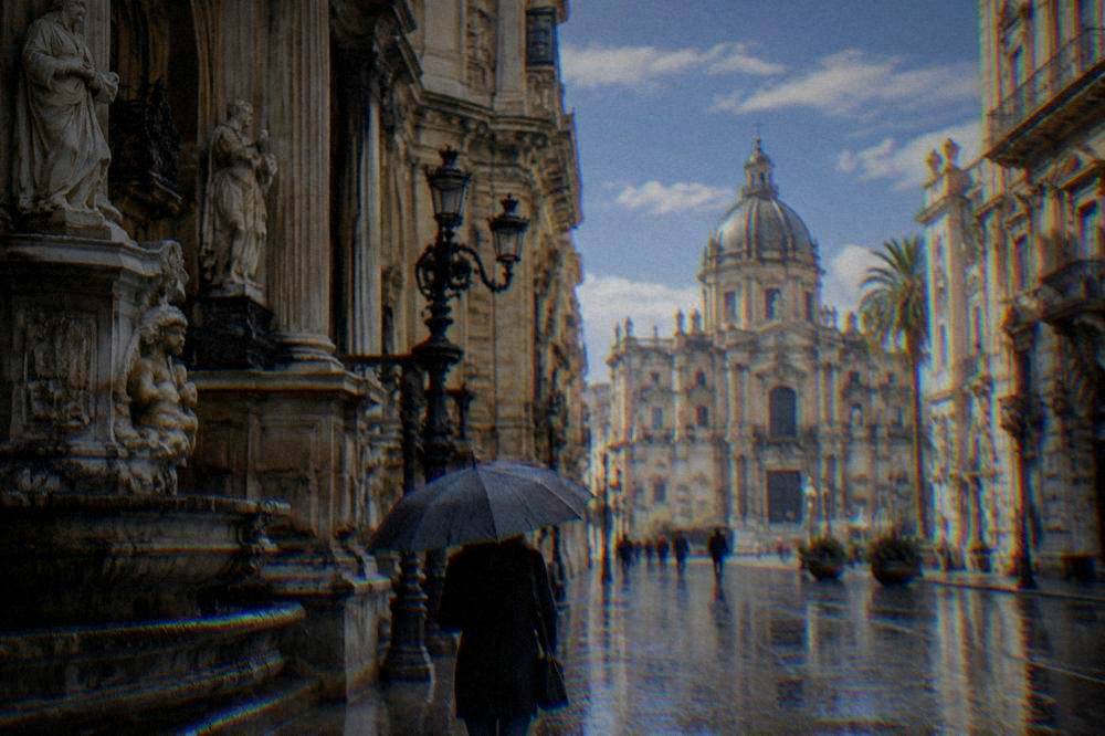 Visitor with an umbrella walking past ornate baroque buildings on a rainy street in Palermo