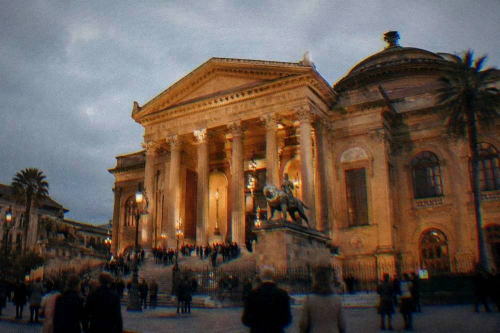 A grand opera house exterior lit at night with people gathered on the steps
